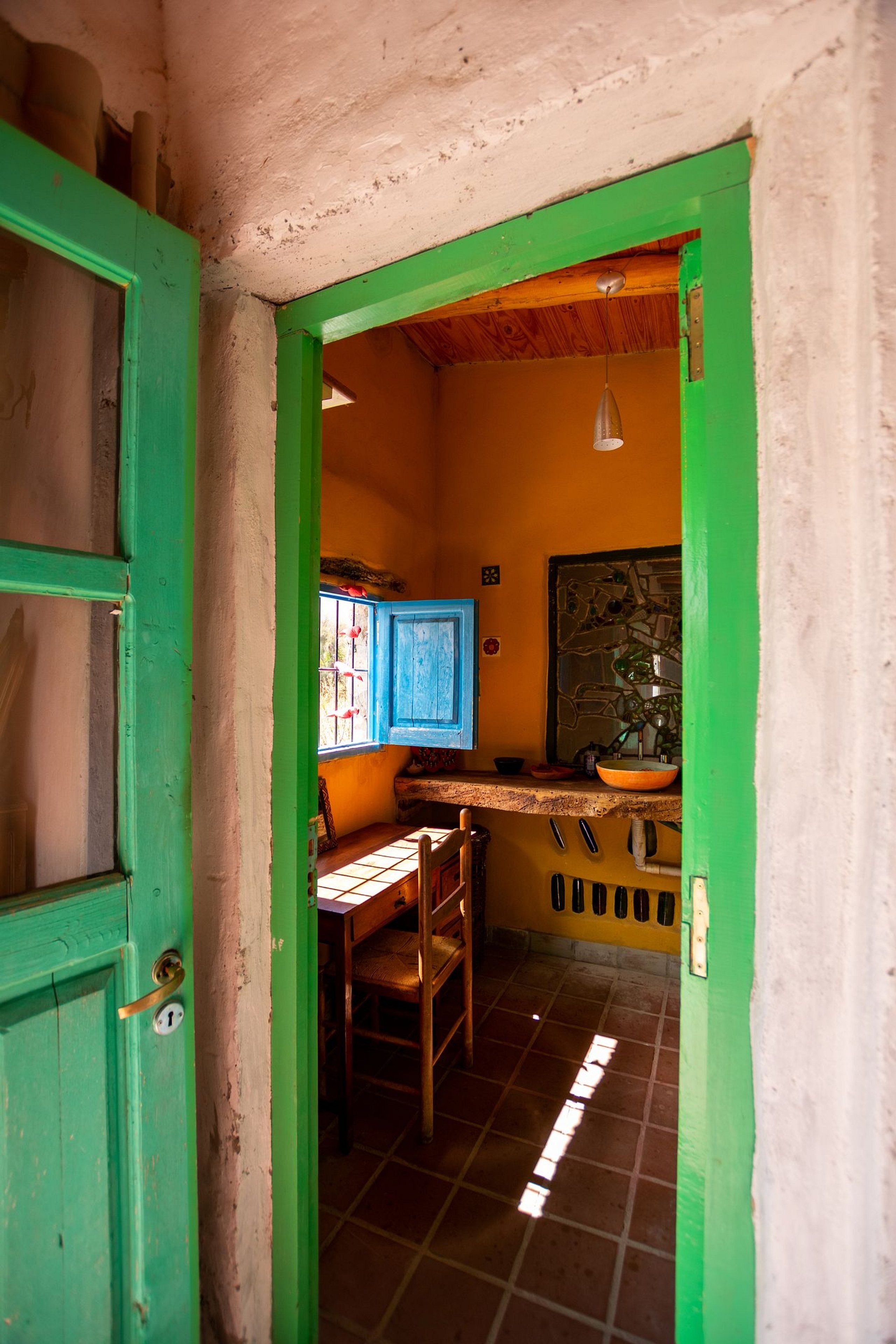 A narrow doorway leading into a room with a tiled floor, a wooden chair, a table, and a window with blue shutters.