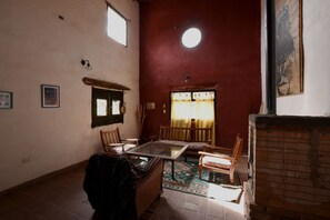 Habitació amb sòl de rajola, una taula rodona, dues cadires, un sofà, una finestra amb cortines i una làmpada fixa a la paret.