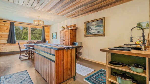 Ein Blockhaus-Interieur mit Holzbalkendecke, einem Essbereich, einer Küche mit Spüle und Herd sowie einem großen Fenster mit Blick auf Grünflächen.