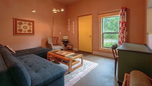 Ein Wohnzimmer mit einer Couch, einem Holztisch, einem Sessel, einem kleinen Tisch mit Lampe und einem Fenster mit Vorhängen.
