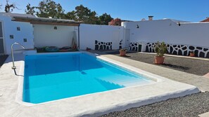 Uma piscina retangular com borda branca, rodeada por uma parede branca com padrão preta e branca, e plantas em vasos.