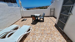Um pátio com uma mesa, cadeiras, espreguiçadeira e vista para o mar.