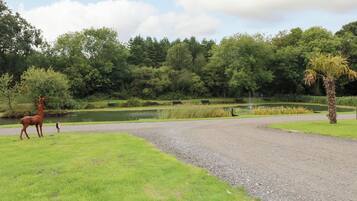 A gravel path leading to a pond with a statue of a deer, surrounded by lush greenery and trees.
