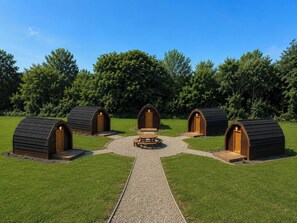 Vier houten hutten met puntige daken, gerangschikt rond een kampvuur op een grindpad, gelegen in een grasrijk gebied met bomen op de achtergrond.