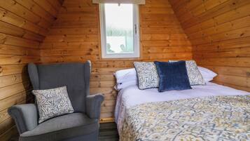 A cozy room with a sloped ceiling, a single bed with patterned bedding, a grey armchair with patterned cushions, and a window with a white blind.