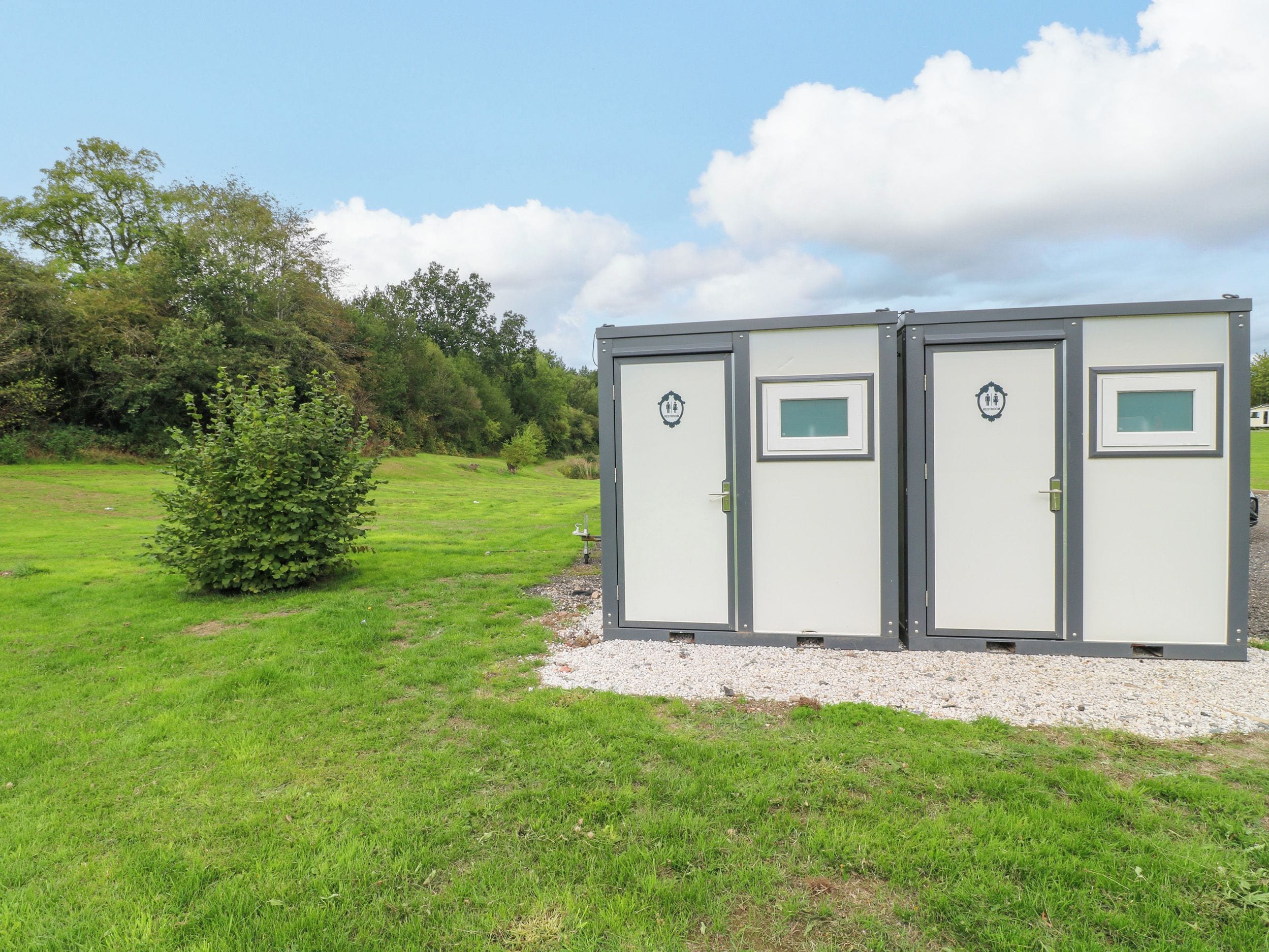 Portable restrooms with a simple design, situated on a grassy field with trees in the background.