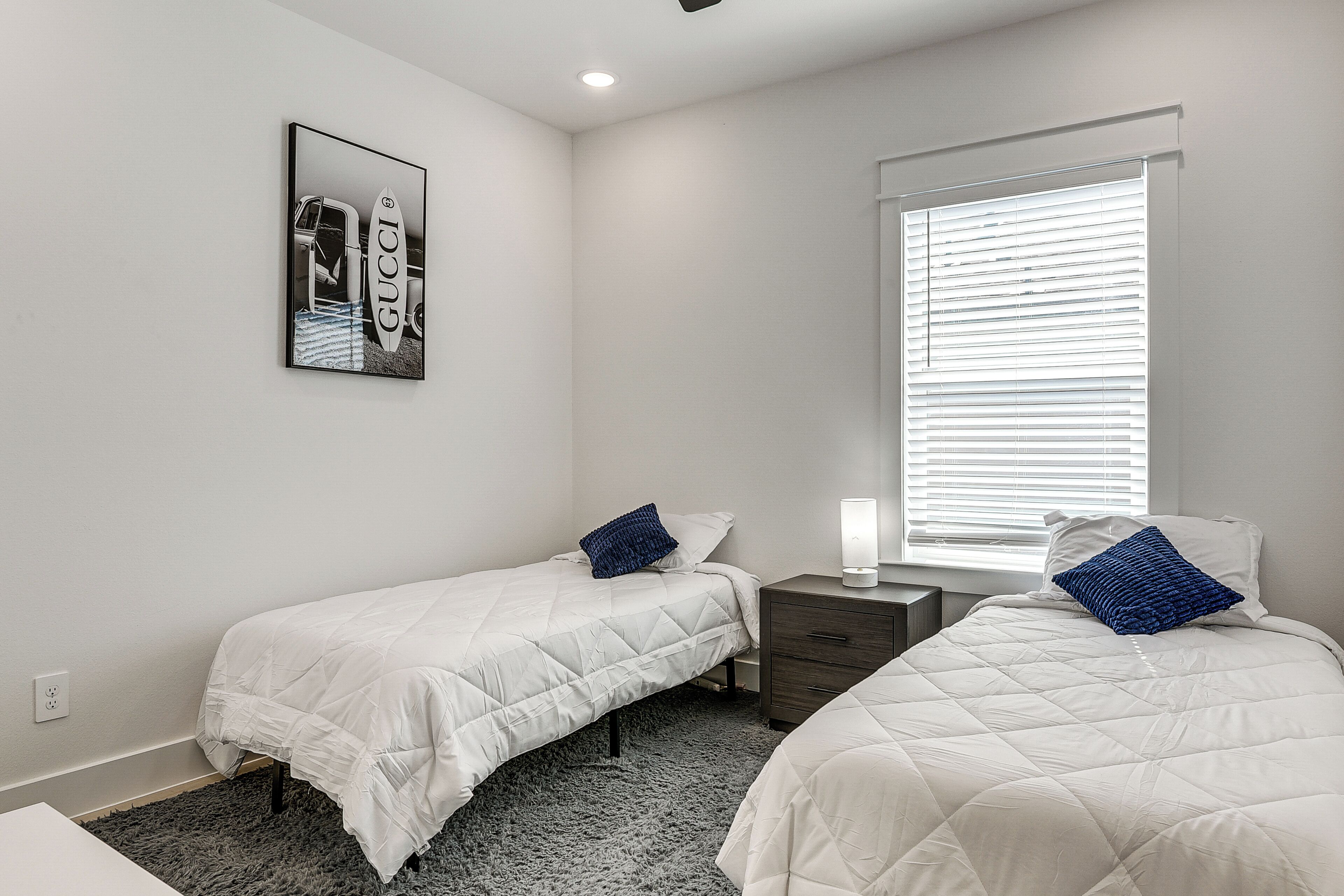 Une chambre avec deux lits, une table de chevet, une commode, une fenêtre avec des stores et un tableau accroché au mur.
