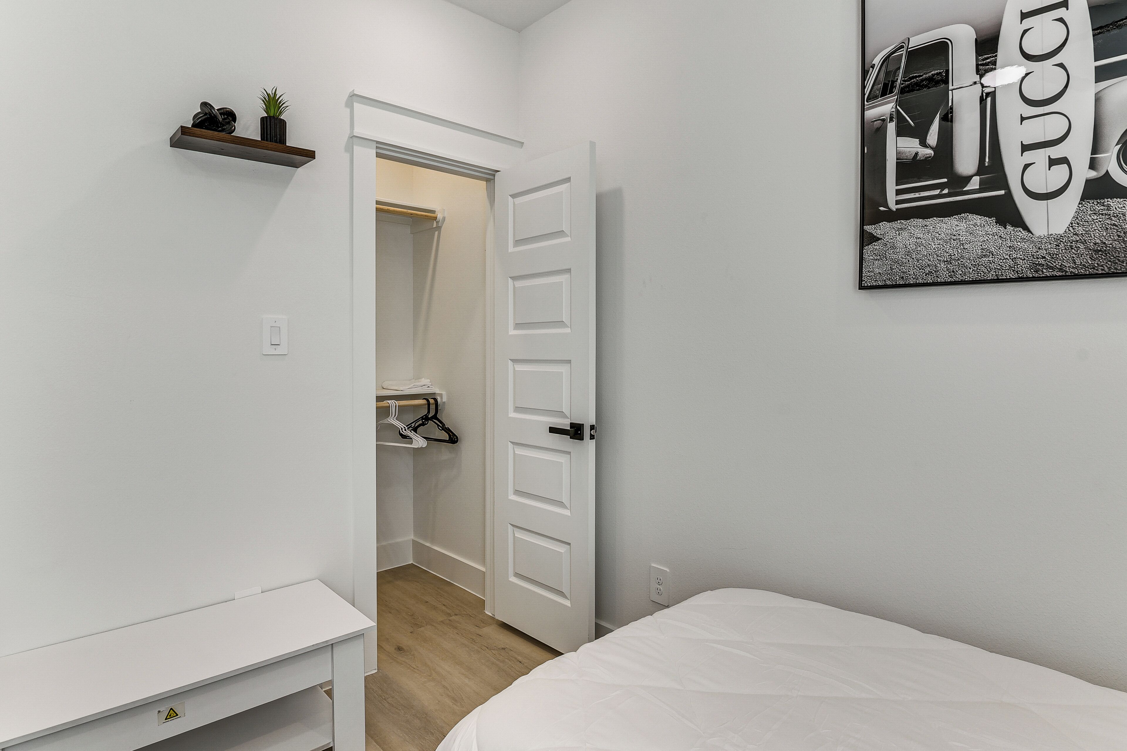 Une chambre à coucher avec un lit, une porte blanche, un mur blanc, une étagère blanche ornée d’une plante et un tableau encadré.