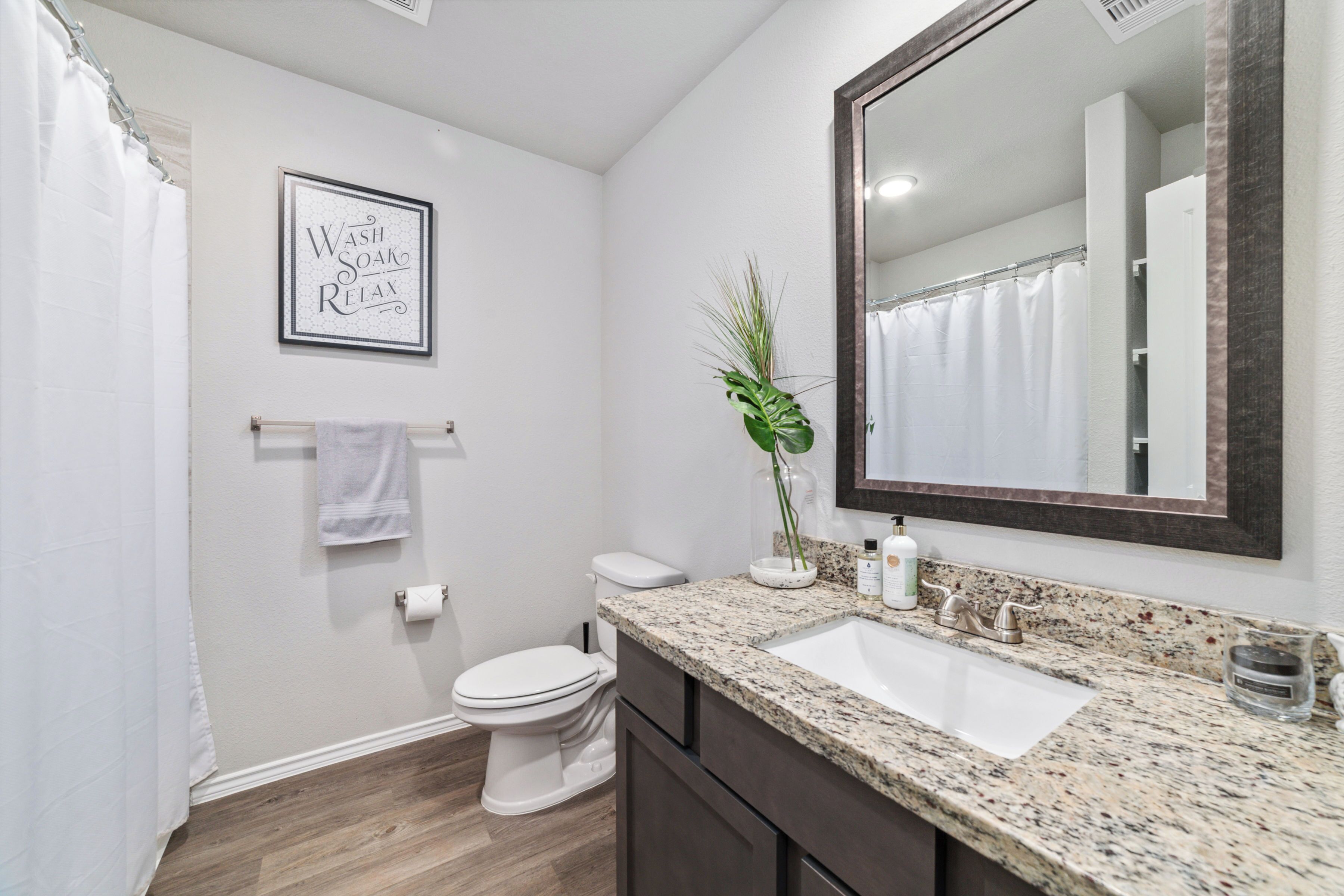 A bathroom with a white toilet, a sink with a granite countertop, a mirror, and a framed sign on the wall.