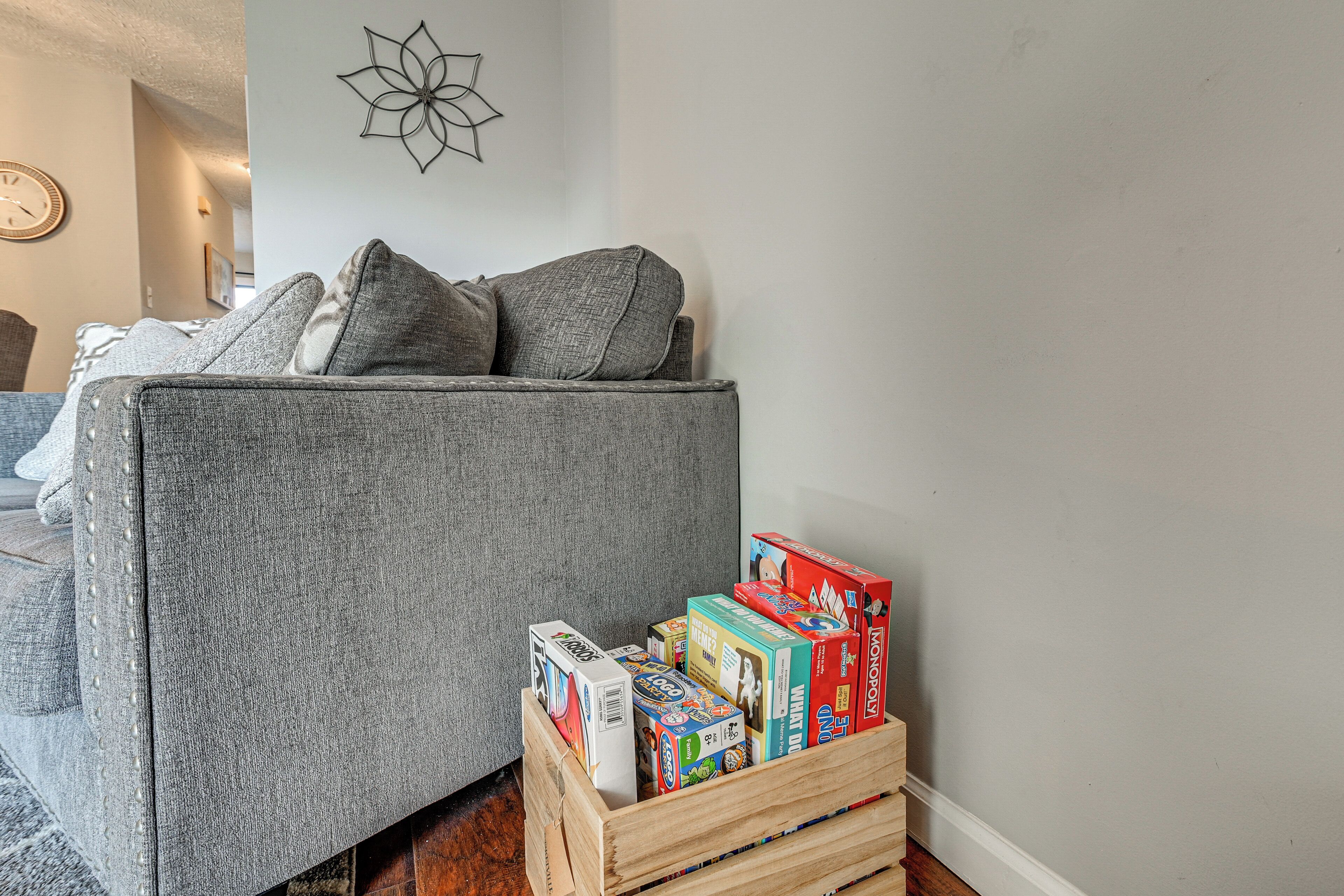 A grey sofa with cushions, a wooden crate with board games, and a wall clock.