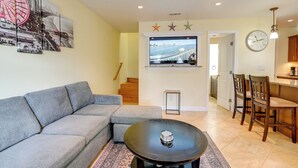 Sala de estar con sofá modular, una mesa de centro, televisor sobre chimenea y comedor con sillas, con cocina al fondo.