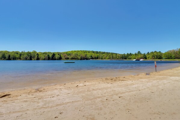 A sandy beach with a calm lake, a green boat, and a forested hill in the background. - Game Room, Deck & Fire Pit! Pocono Mtn House (Blakeslee)