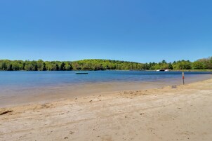 Uma praia arenosa com um lago calmo, um barco verde e uma colina arborizada ao fundo.