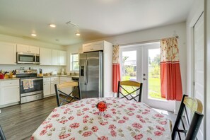 A kitchen with white cabinets, a microwave, a refrigerator, and a dining table with a floral tablecloth. - Serene Punta Gorda Home Near the Peace River (Punta Gorda)