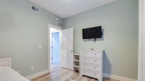 Un dormitorio con cama, cómoda, televisor montado en la pared y puerta que da a otra habitación.