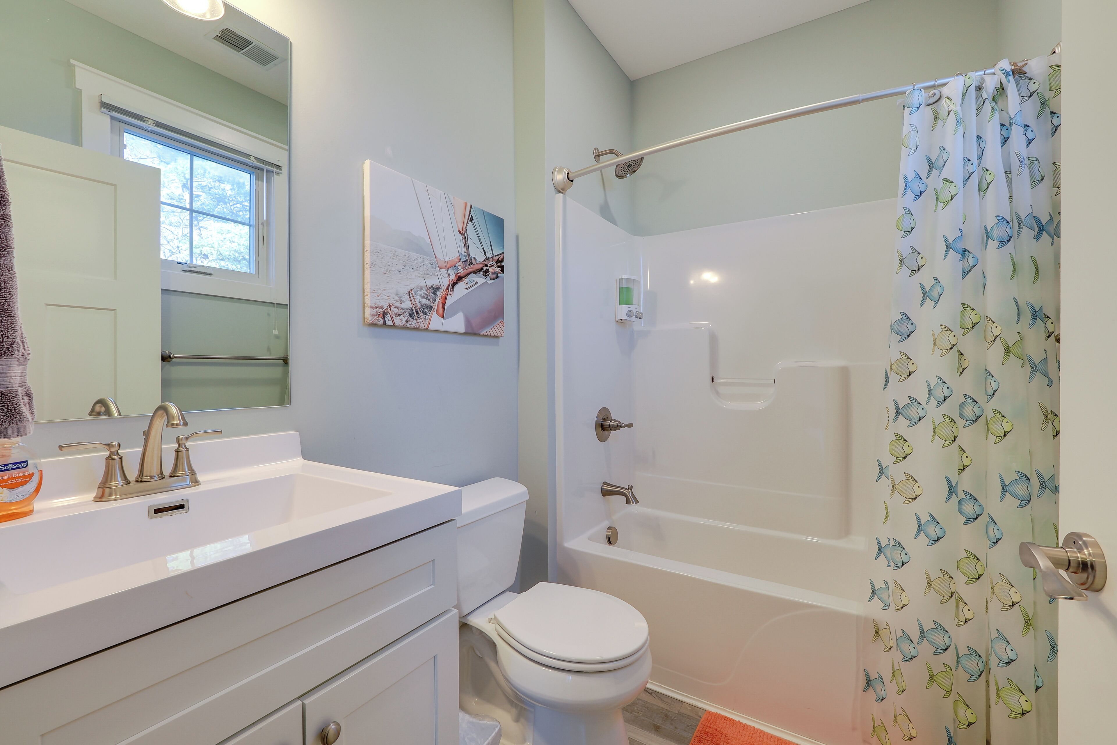 Une salle de bain avec un lavabo, des toilettes et une baignoire, tous de couleur blanche. Il y a une fenêtre au-dessus du lavabo et un rideau de douche orné de motifs de poissons.