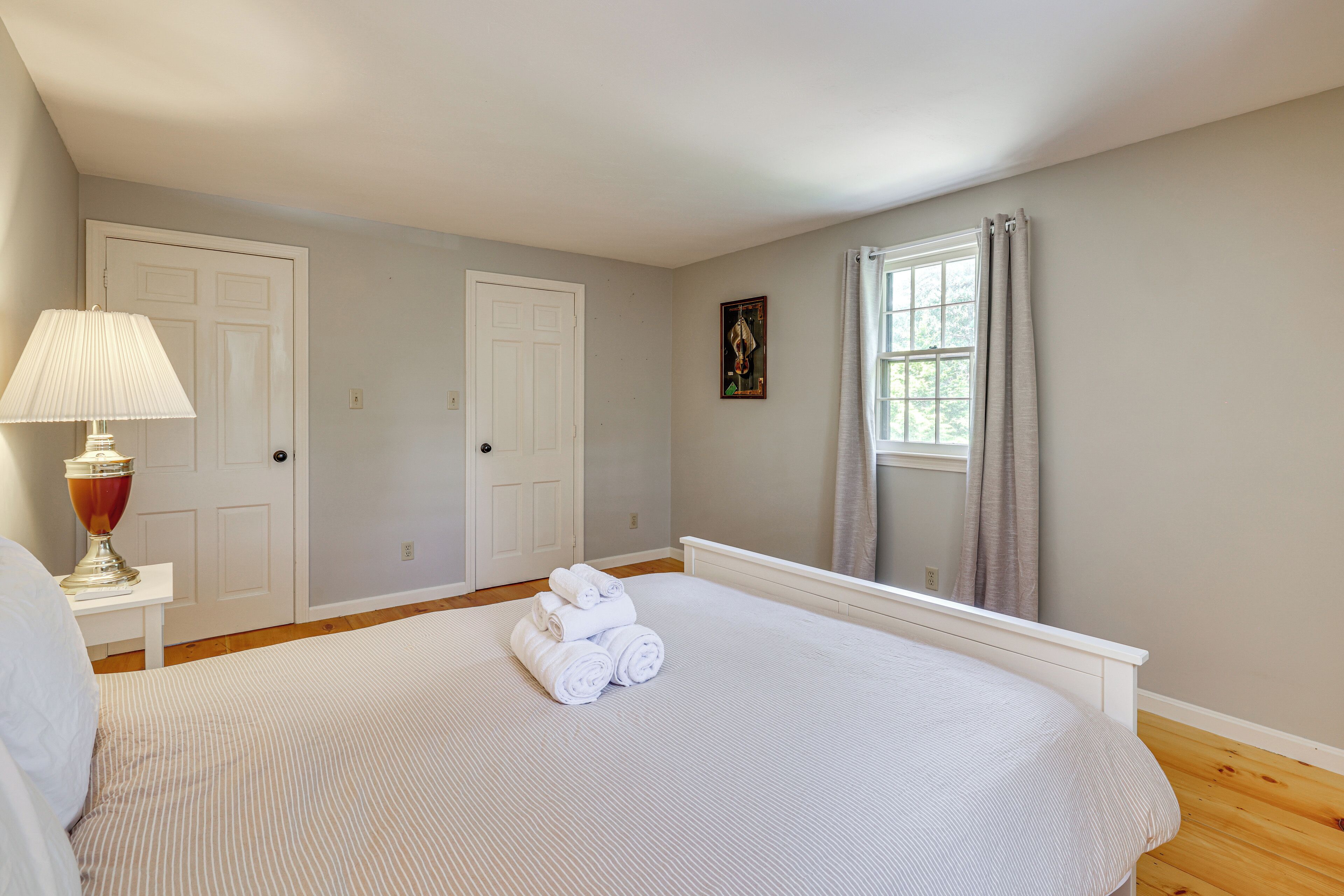 Un dormitorio con cama, una mesita de noche con lámpara, dos puertas, una ventana con cortinas y un cuadro colgado en la pared.