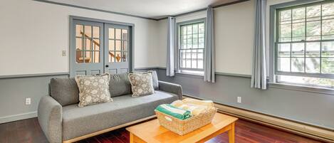 A living room with a gray sofa, two windows, a wooden table, and a basket of towels.