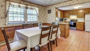 A kitchen with wooden cabinets, a dining table with chairs, a large window with curtains, and a tiled floor. - Classic Cape House 5M to Nauset Beach (Orleans)