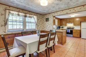 A kitchen with wooden cabinets, a dining table with chairs, a large window with curtains, and a tiled floor. - Classic Cape House 5M to Nauset Beach (Orleans)