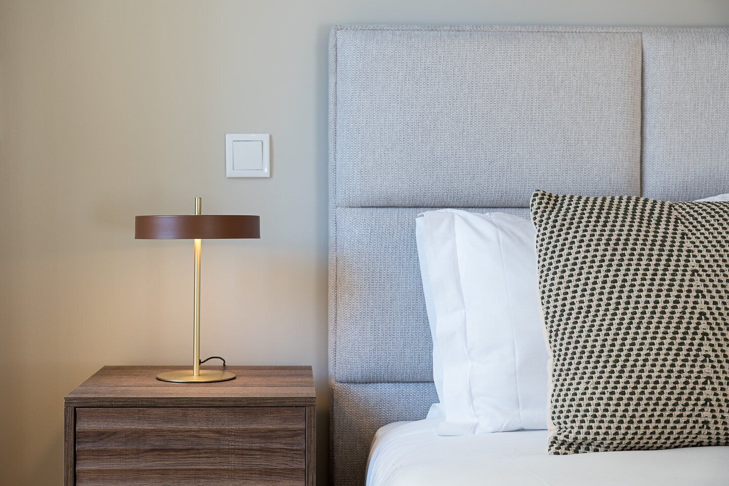 A neatly made bed with white and patterned pillows, a bedside lamp, and a wooden nightstand.