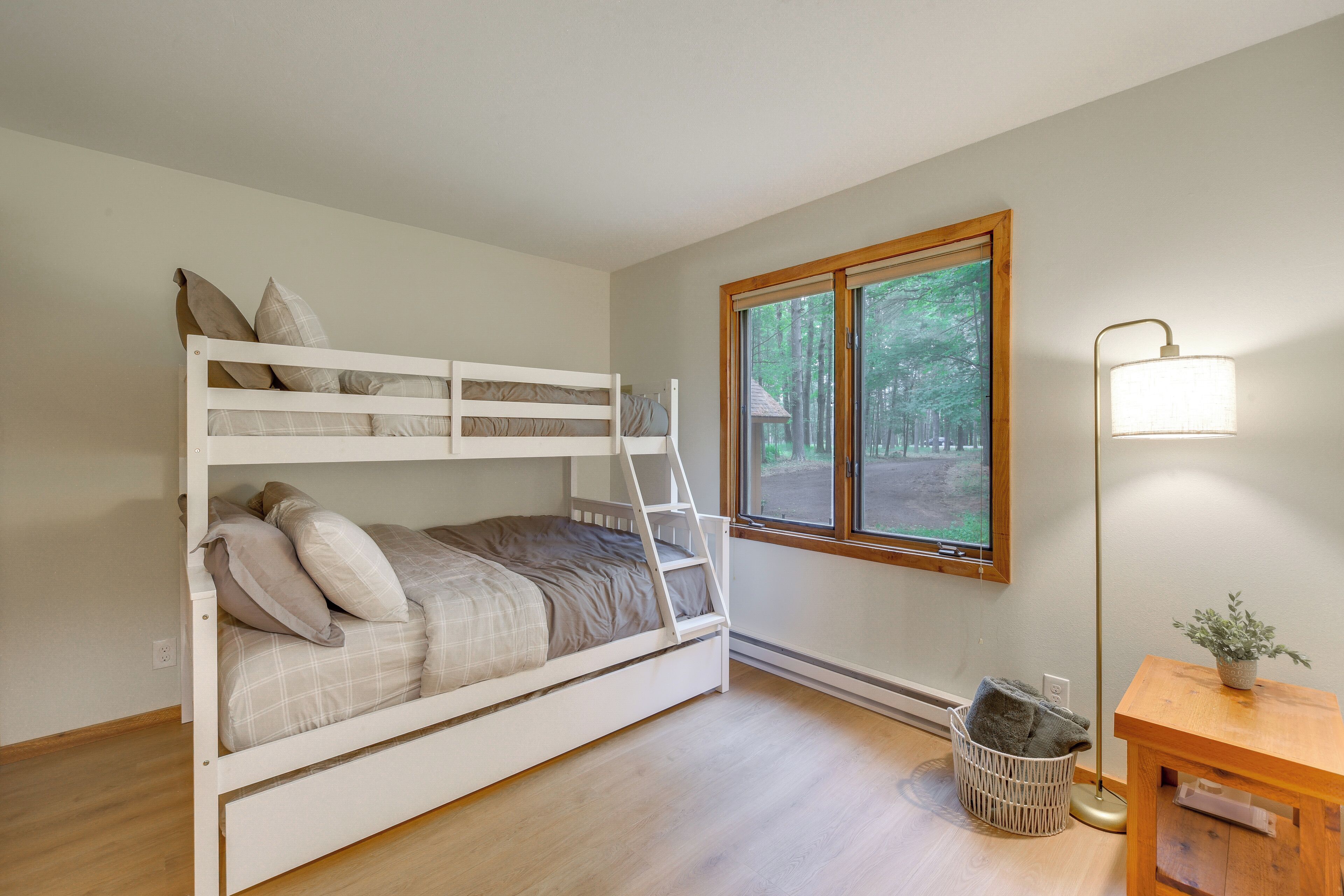 A bunk bed with a ladder, a window with a view of trees, a floor lamp, and a small table with a plant.