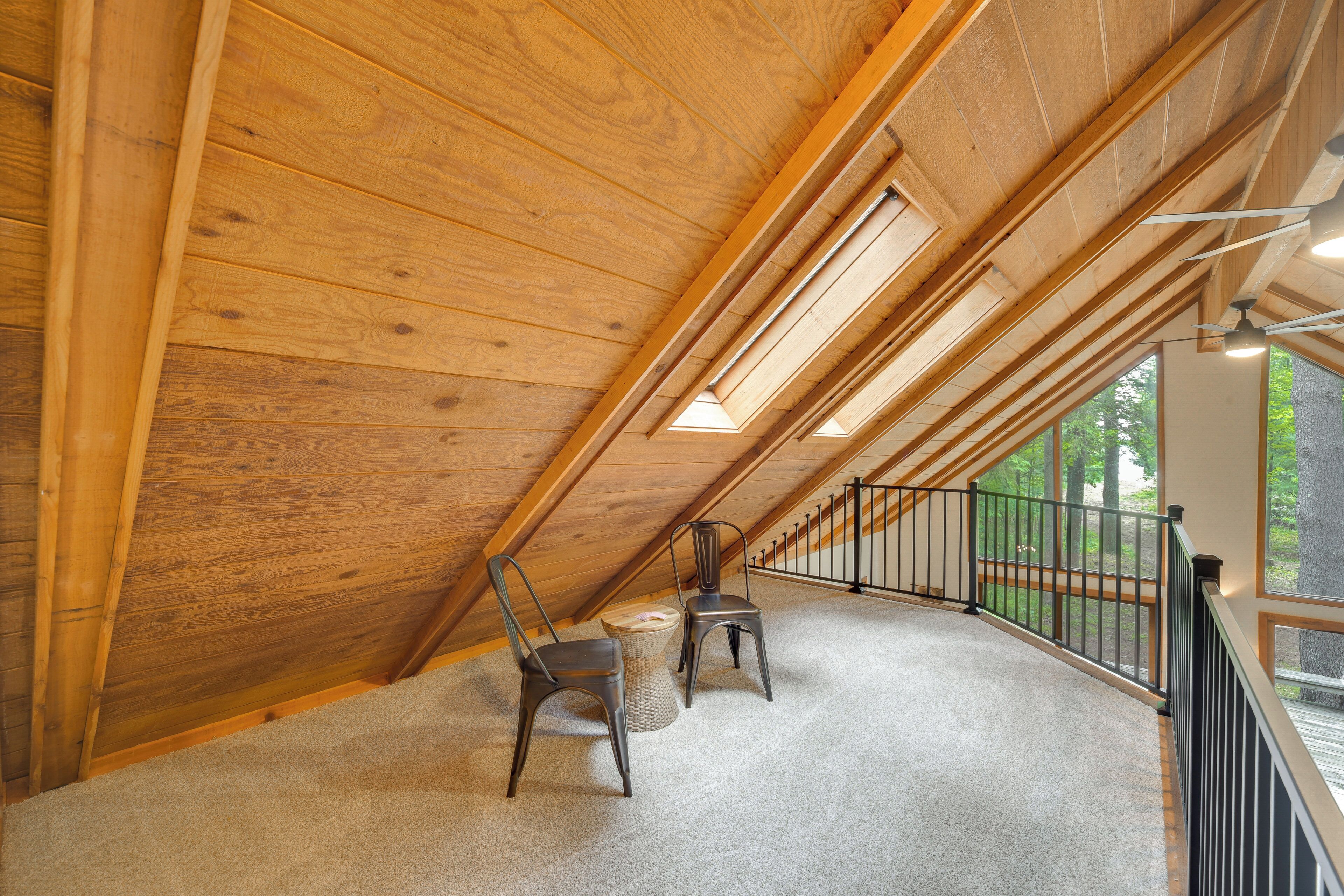 A wooden attic room with a sloped ceiling, two chairs, a small round table, and a view of greenery outside.