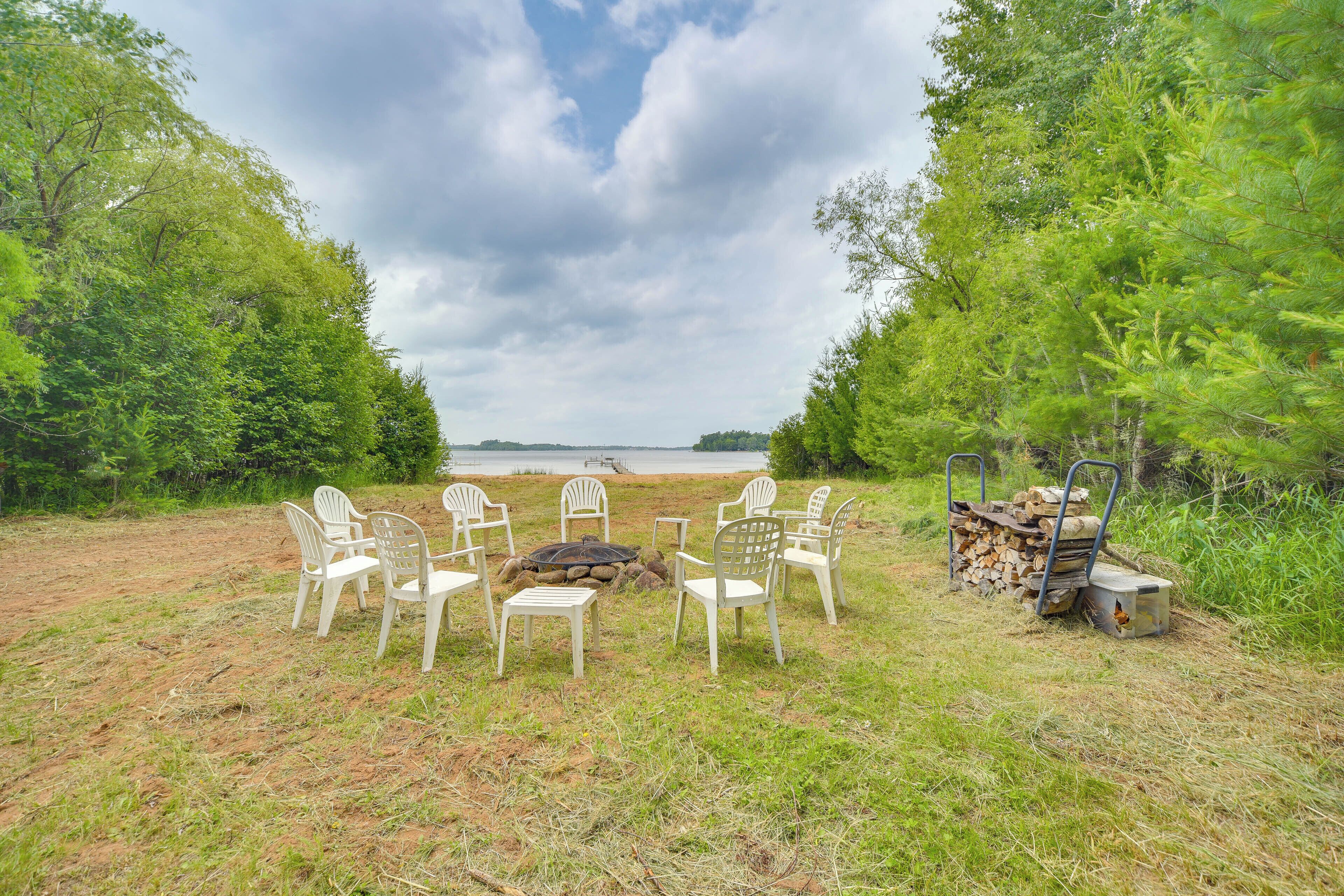 A campsite with white plastic chairs arranged around a central fire pit, surrounded by trees and a body of water in the background.