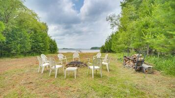 A campsite with white plastic chairs arranged around a central fire pit, surrounded by trees and a body of water in the background.