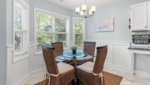 Uma área de jantar com mesa de vidro, cadeiras de vime, lustre e vista para o verde através de grandes janelas.
