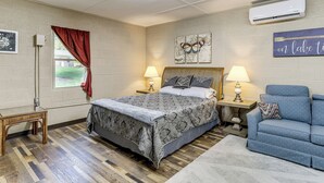 A bedroom with a bed, two bedside lamps, a blue sofa, a wooden floor, a window with a red curtain, and a wall-mounted air conditioner. - Studio w/ Greers Ferry Lake Access & Resort Perks! (Clinton)