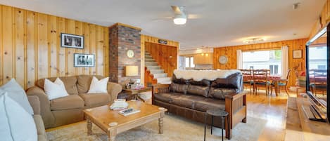 Un salon chaleureux avec un canapé en cuir marron, une table basse en bois et un divan marron. Un escalier mène à un niveau supérieur, et on y trouve un coin repas avec des chaises et une table en bois.