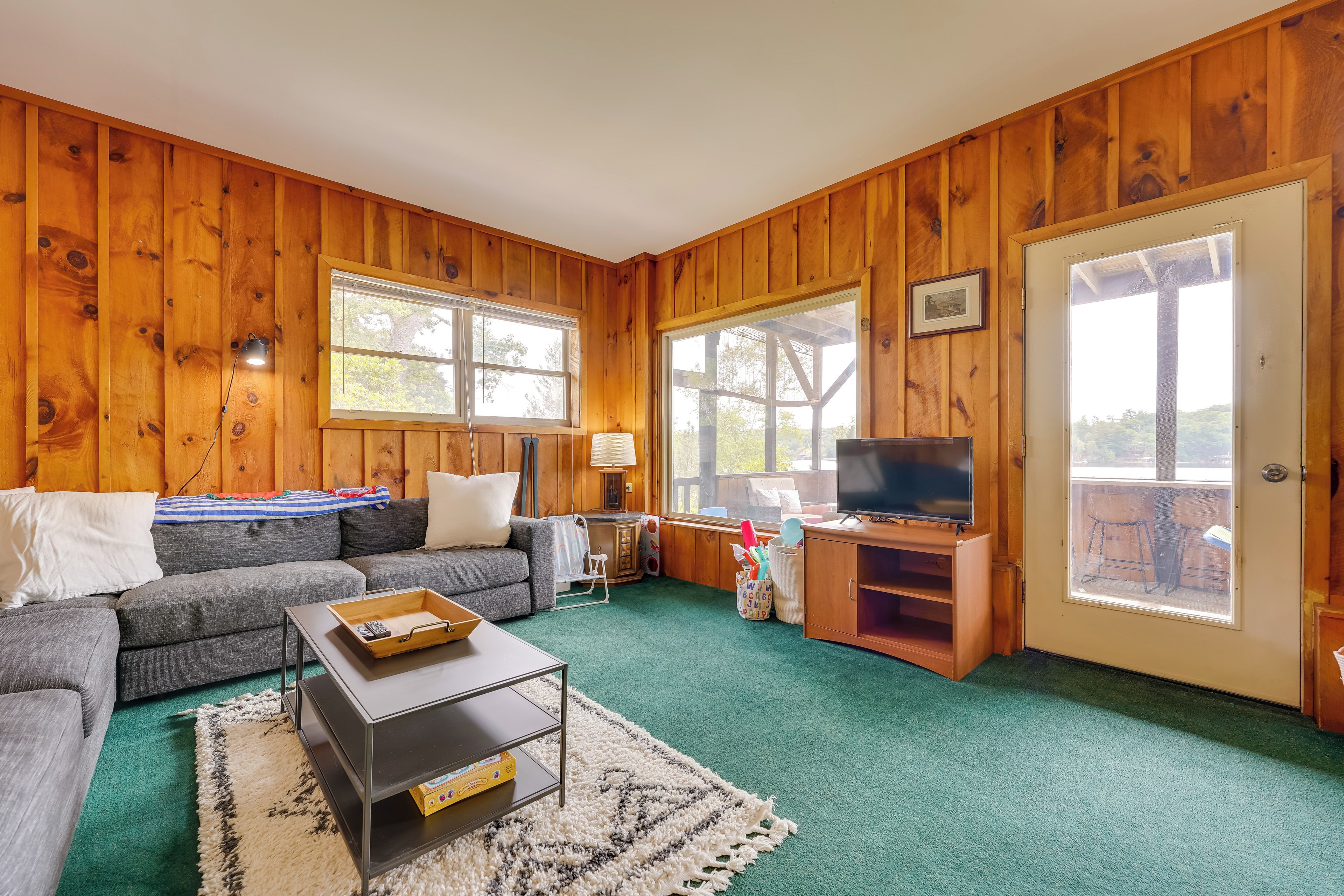 Una habitación con paneles de madera, un sofá, un televisor sobre un mueble de madera, una mesita y una ventana.