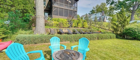 Ein modernes, zweistöckiges Haus mit einer großen Terrasse, umgeben von üppiger Vegetation und einer Feuerstelle mit Sitzgelegenheiten.