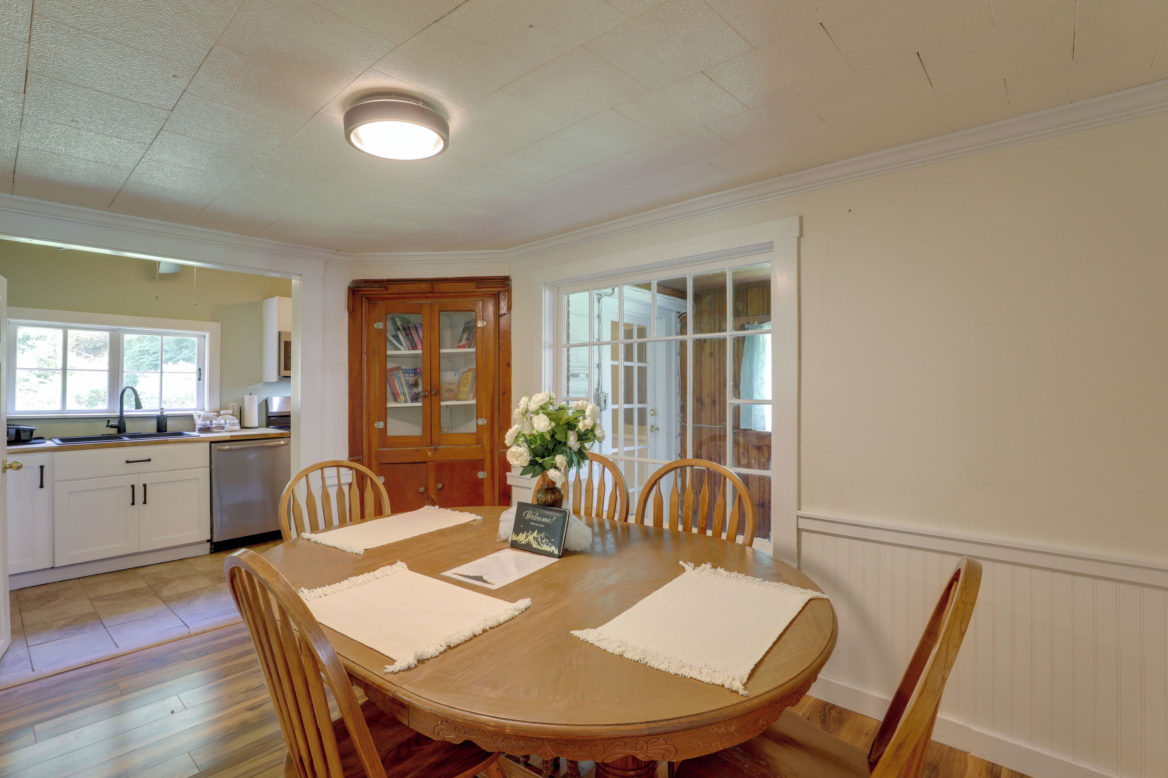 A dining room with a wooden table, chairs, a vase with flowers, and a view of the kitchen and dining area.