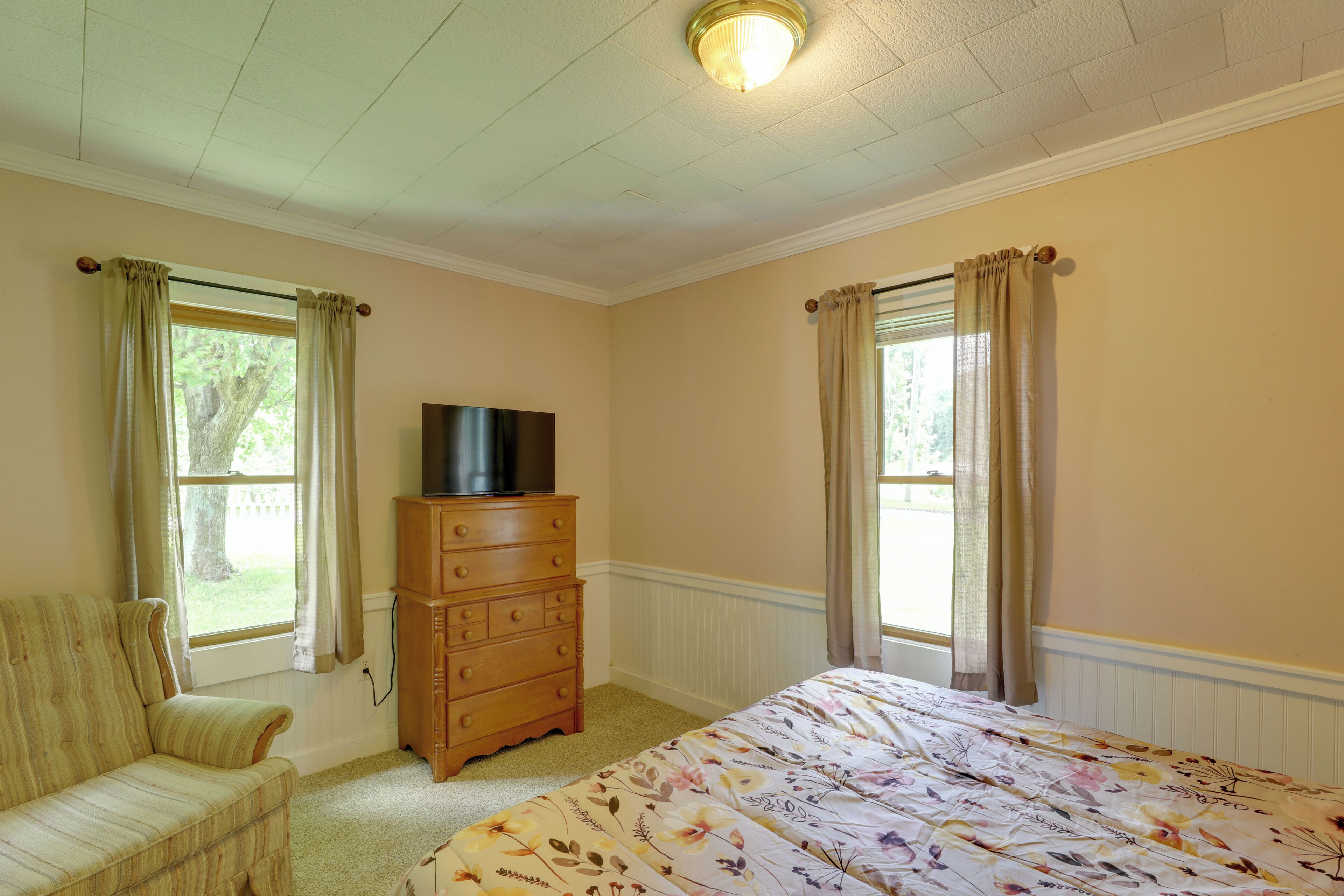 A bedroom with a bed, a dresser, a television on top, and two windows with curtains.