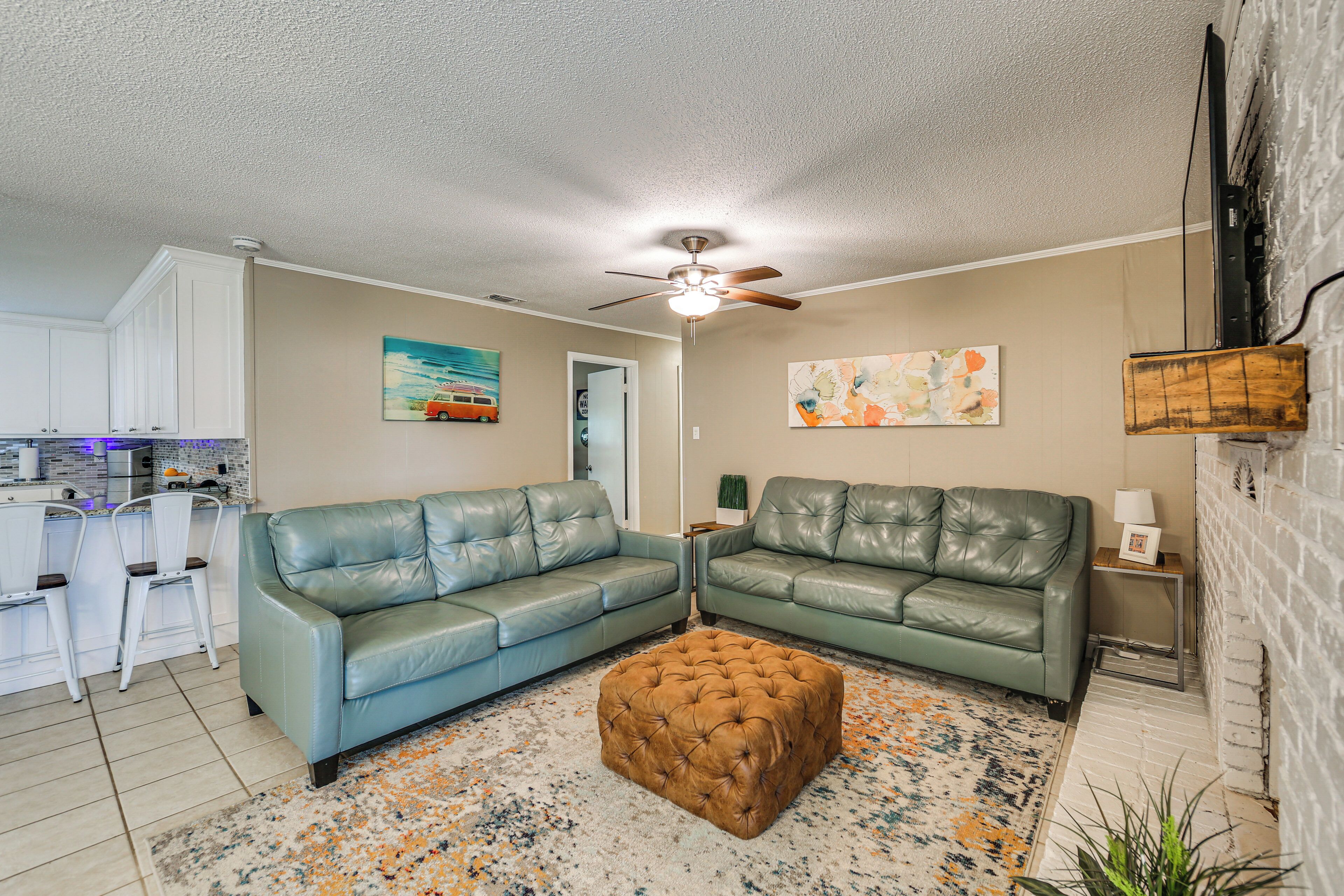 A living room with two green leather sofas, a tufted ottoman, a ceiling fan, and a kitchen area with white cabinets and bar stools.