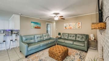 A living room with two green leather sofas, a tufted ottoman, a ceiling fan, and a kitchen area with white cabinets and bar stools.