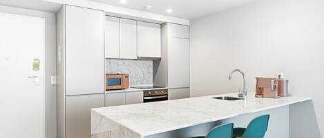 A modern kitchen with white cabinetry, a marble countertop, a built-in oven, a microwave, and a sink. There are two teal chairs at the island.