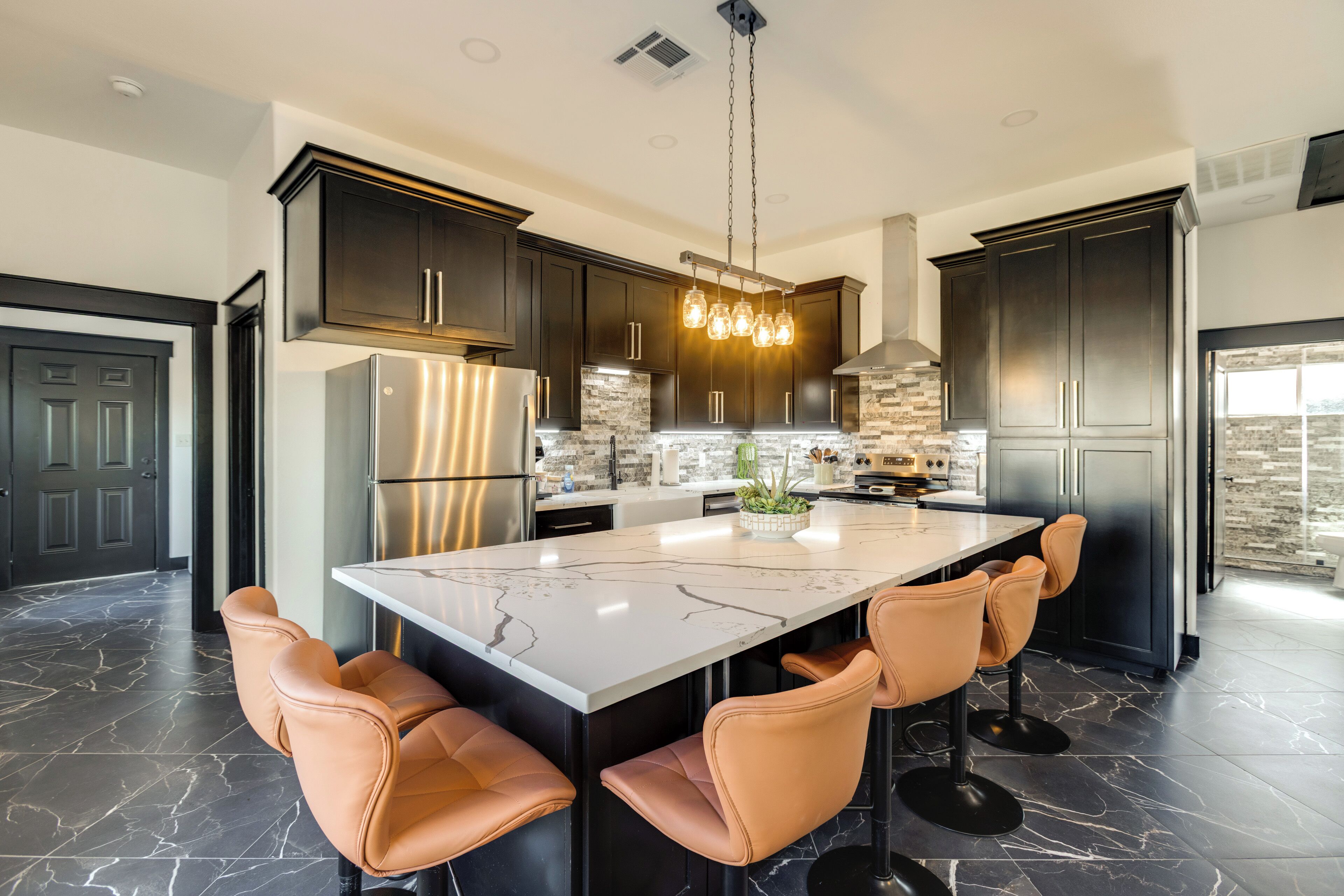 A modern kitchen with dark cabinets, a large island with bar stools, stainless steel appliances, and a hanging light fixture.
