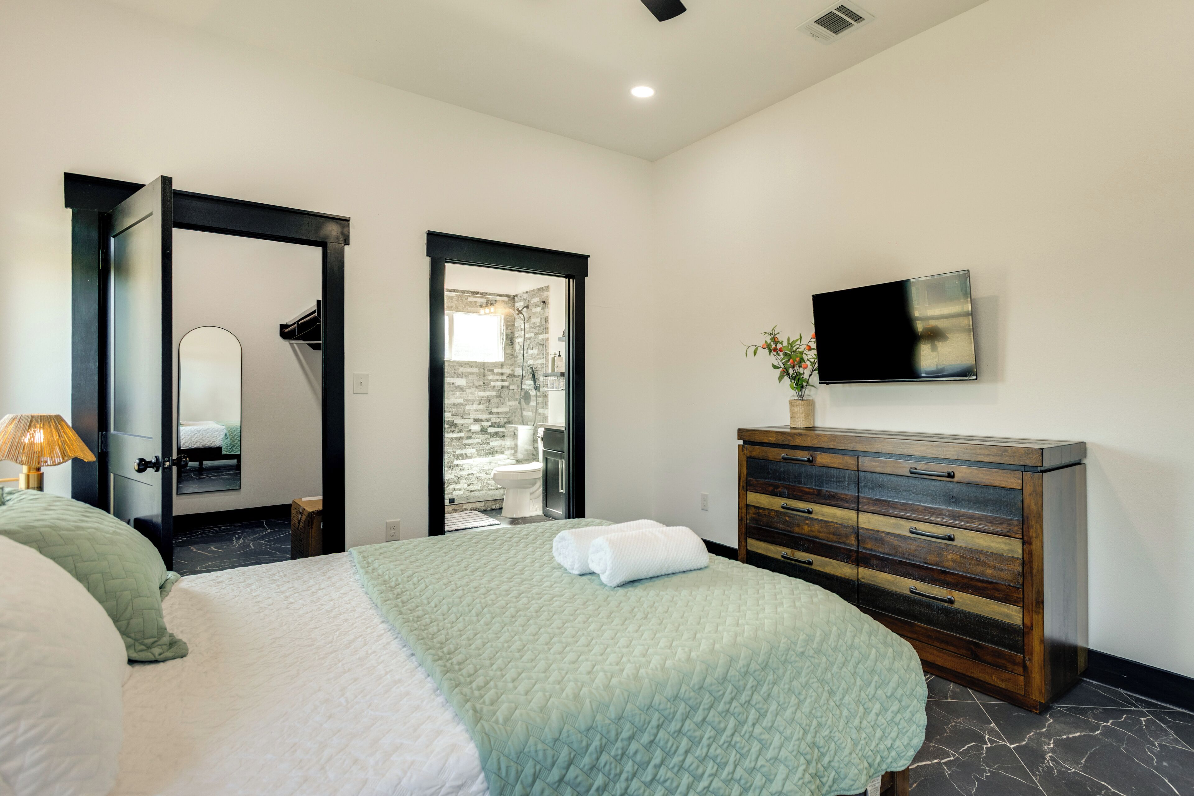 A bedroom with a bed, a dresser, a TV, and a bathroom visible through an open door.