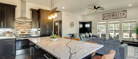 A modern kitchen with dark cabinets, a marble island, and a stone backsplash. There is a dining area with a gray sofa and a ceiling fan.