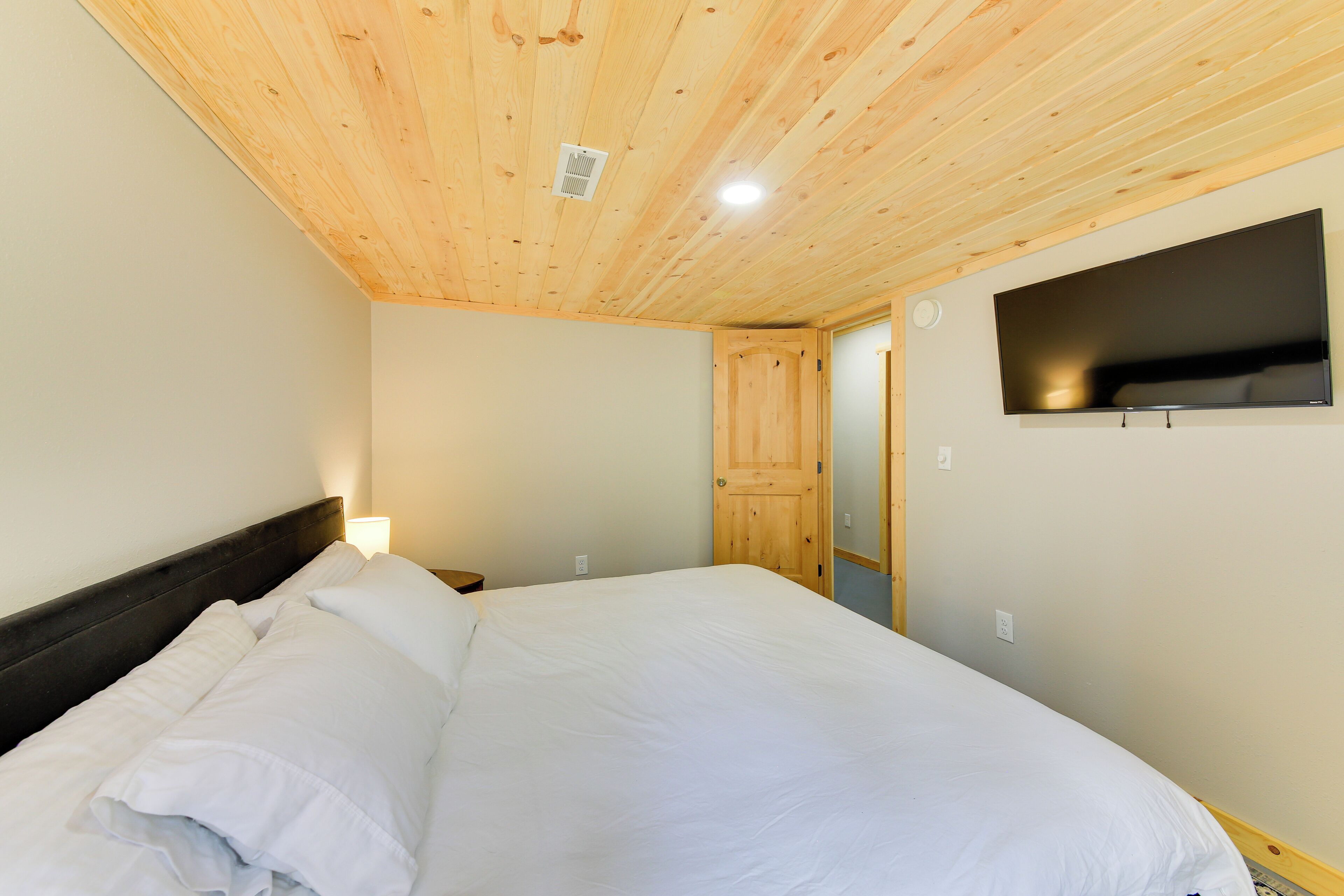Een slaapkamer met een houten plafond, een bed met wit beddengoed, een tv aan de muur en een deur naar een andere kamer.