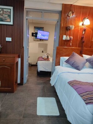 Ein Hotelzimmer mit einer Holzwand, einem Fernseher, einem Bett mit weißer und violetter Bettdecke, einem kleinen Tisch mit einer Vase sowie einem Holzkabinett.
