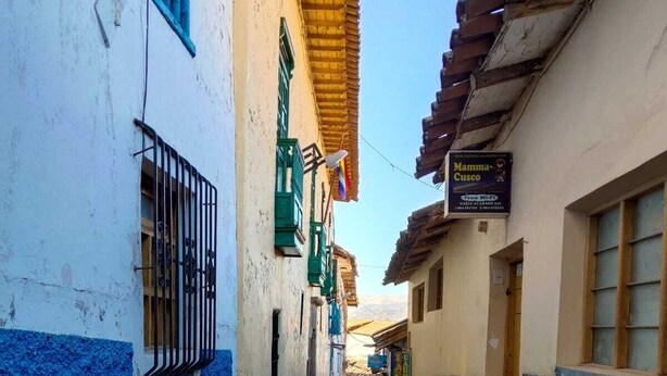 En smal gränd med kullerstenar, med byggnader på båda sidor, krukväxter och en skylt med texten ”Mamina Cusco”.