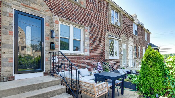 Une maison en briques avec une porte d’entrée, des marches, un petit espace extérieur aménagé avec une table et des chaises, et un arbuste vert.