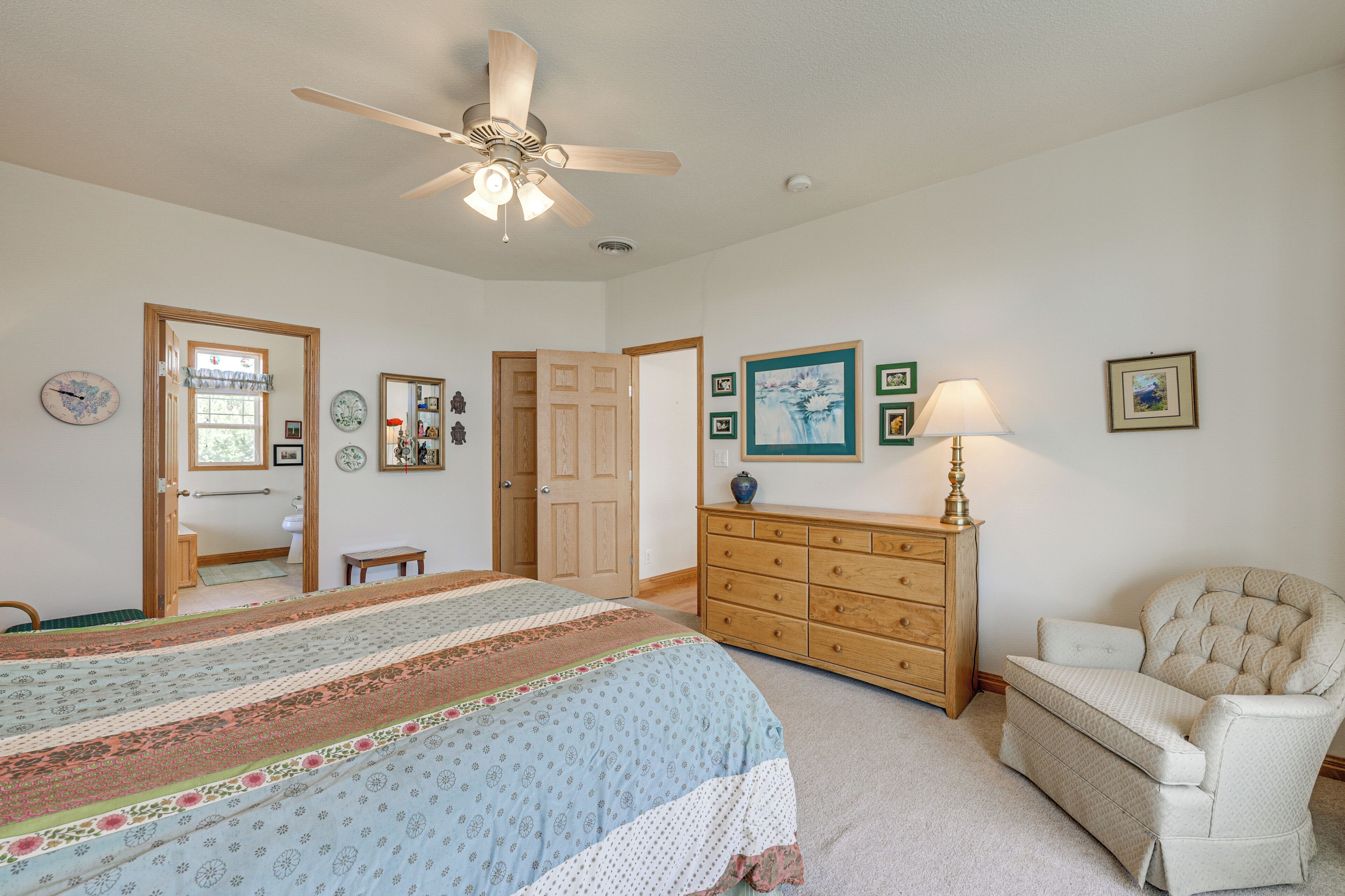 A bedroom with a bed, a dresser, a chair, a ceiling fan, and a door leading to another room.