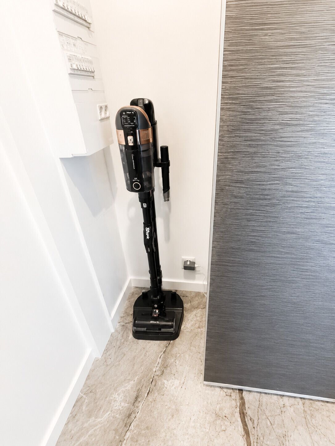 A vacuum cleaner in a corner of a room with a tiled floor and a metallic panel on the wall.