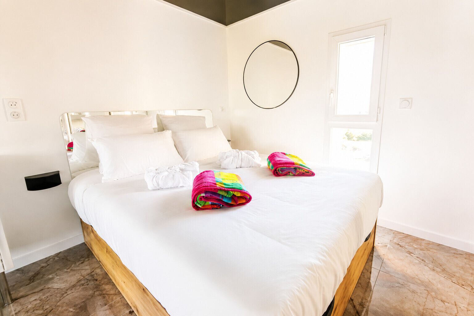 A hotel room with a bed, white linens, a circular mirror, a window, and a marble floor.