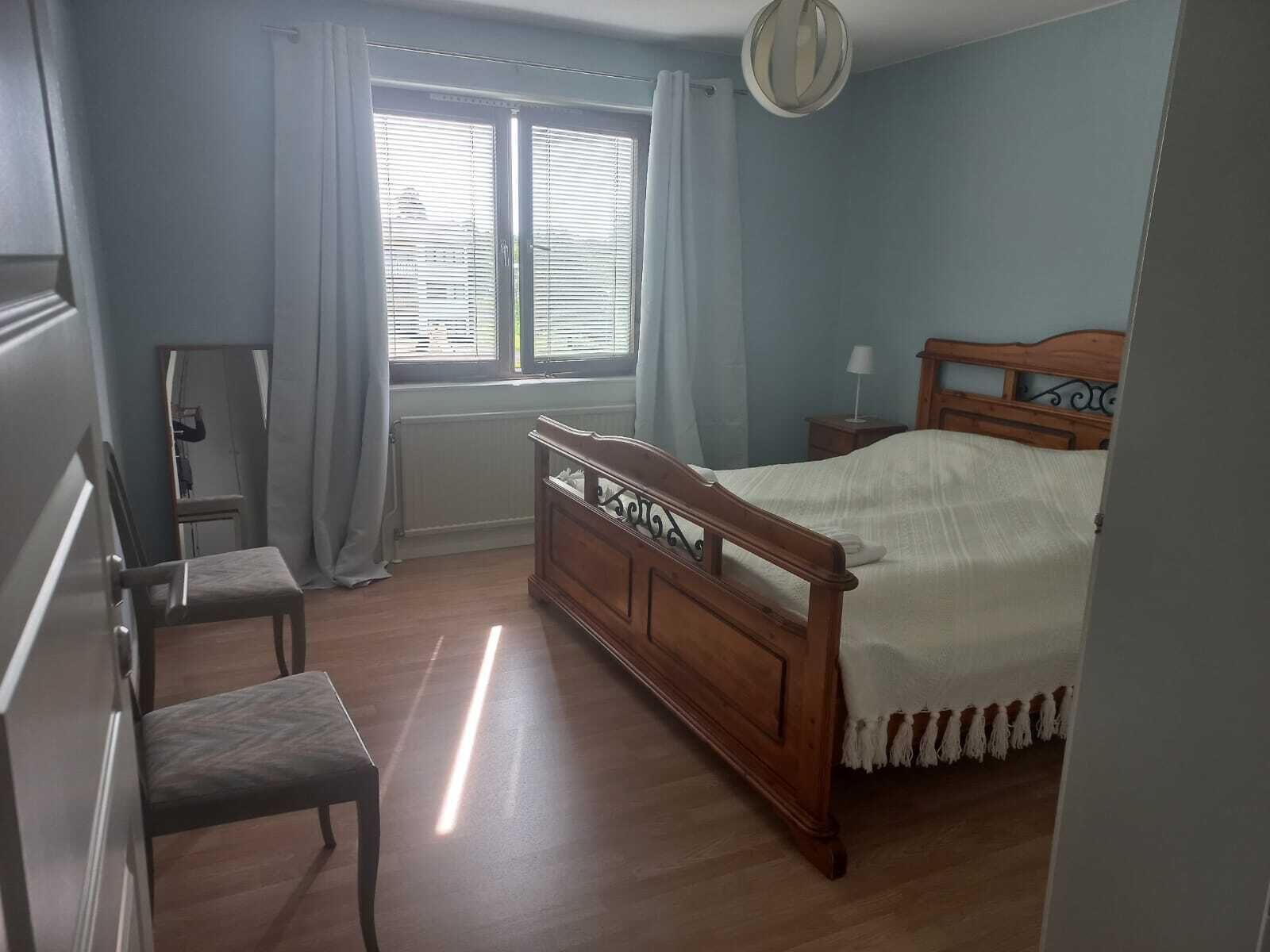 Un dormitorio con cama de madera, una silla, una mesita de noche, un espejo y una ventana con persianas.